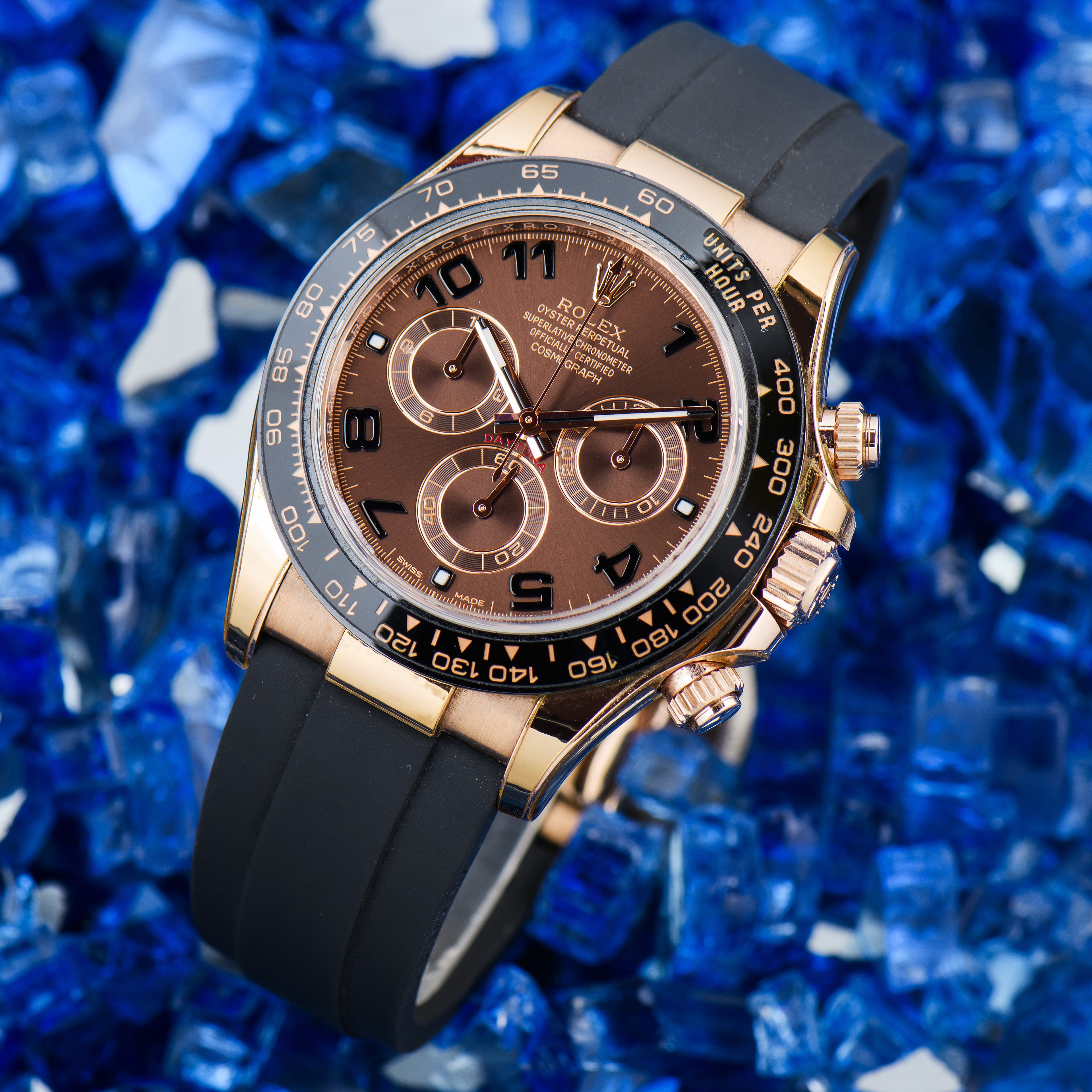 A genuine Rolex Cosmograph Daytona in Everose gold with a chocolate dial, shown dial-forward on a blue crushed-glass background